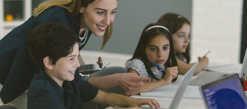 Kids at coding class. They are sitting by the desk and using laptop, coding. Selective focus on female teacher helping to male student with code.