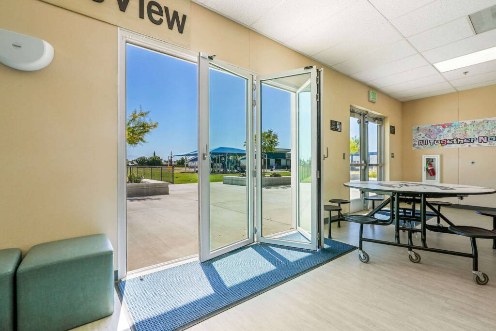 View from inside a building looking out through open glass doors toward a patio area with trees and a blue-roofed structure in the distance.