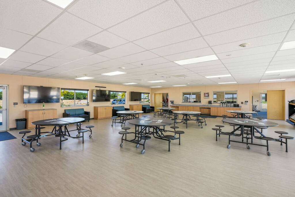Large multipurpose room with round mobile tables and stools, wall-mounted TVs, windows and light wood flooring.