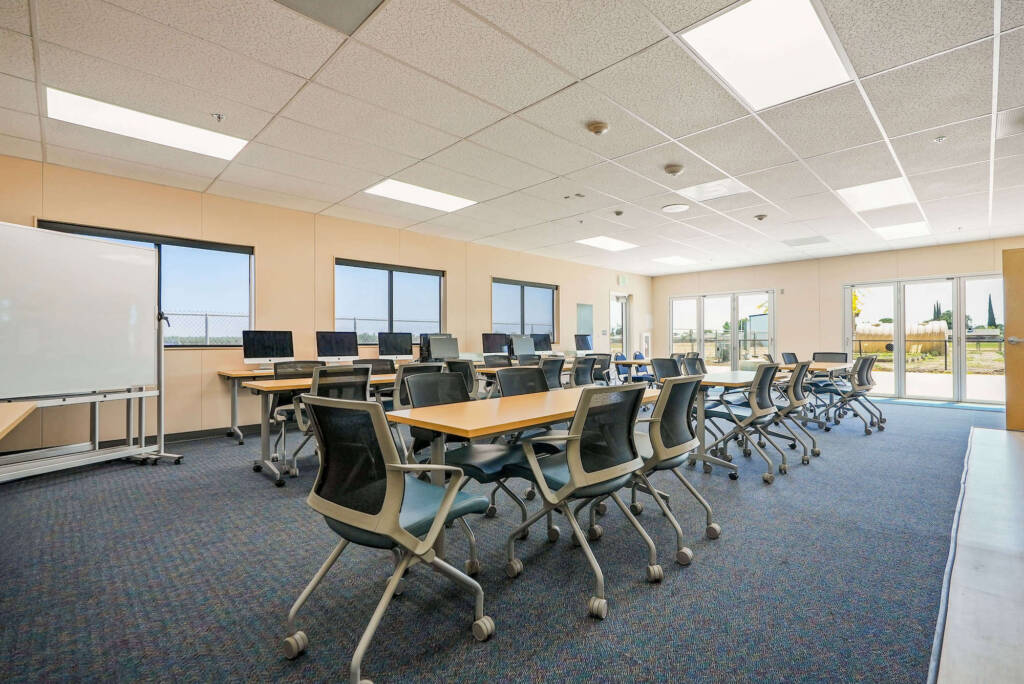 Classroom with rolling chairs, rectangular tables, carpeted floor and large windows and glass doors letting in natural light.