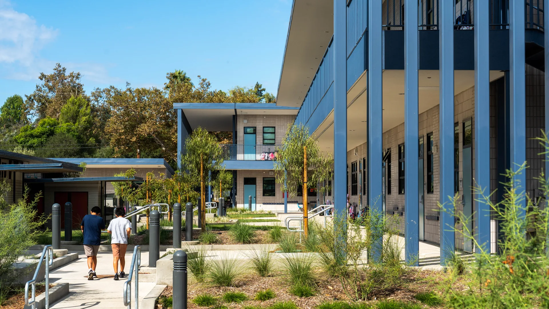 Two people walking along a landscaped path beside a completed two-story AMS modular school building